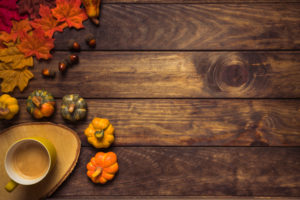 Wooden background with fall leaves and gourds and a mug of coffee in the lower left hand corner. The words